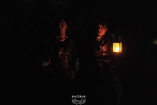 Due personaggi camminano di notte alla luce di una lanterna, Exclave settembre 2026