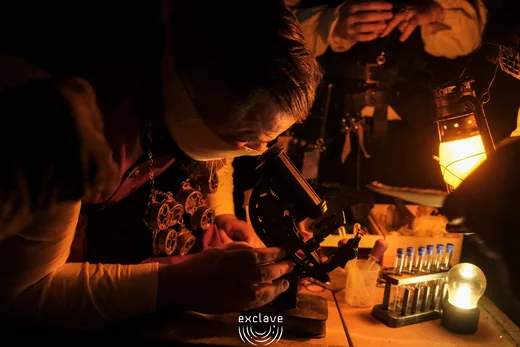 Un personaggio prende appunti al banco alchemico illuminato da una lanterna, Exclave settembre 2026